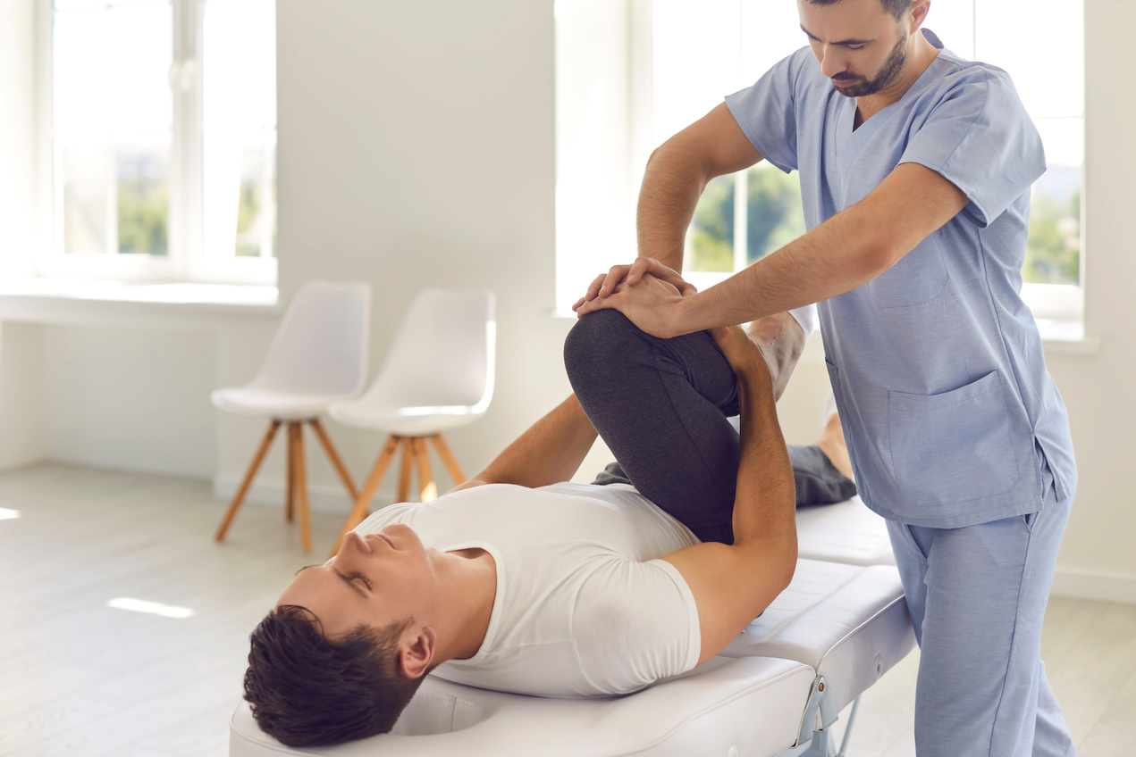 Serious Doctor Helping Male Athlete Do Physical Exercise during Rehabilitation after Leg Injury